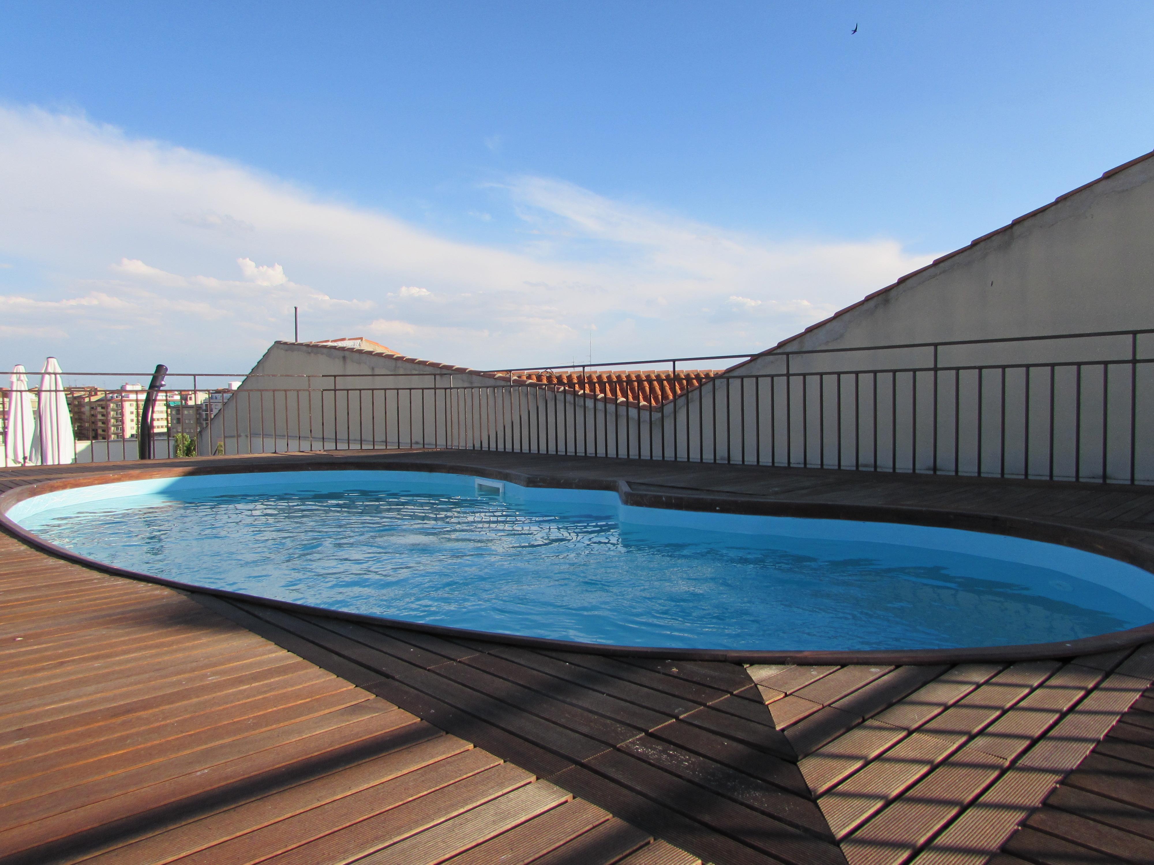 Hotel Alameda Palace Саламанка Экстерьер фото The photo shows a rooftop space featuring a modern, oval-shaped swimming pool. The pool has a light blue color and is surrounded by a wooden deck. In the background, there are slanted roofs, and the sky is clear with a few clouds. The overall setting